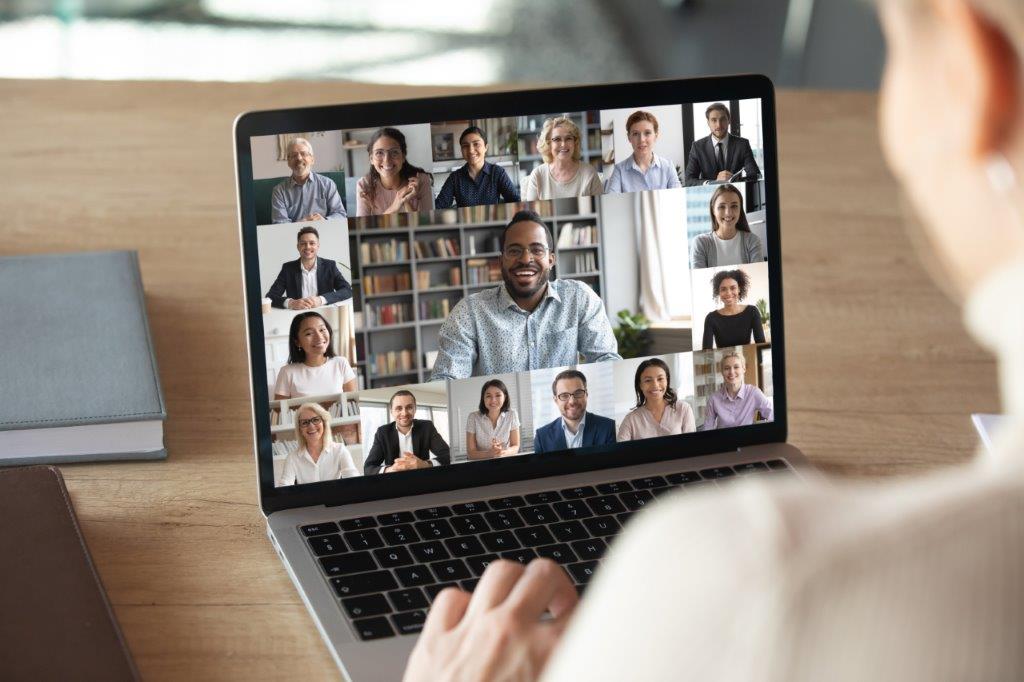 Person blickt über einen Monitor in eine Videokonferenz mit verschiednen Menschen.