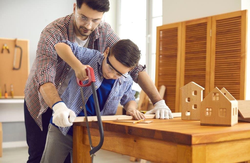 Ein Junge mit Schutzbrille sägt ein Holz, das von einem Mann gehalten wird.